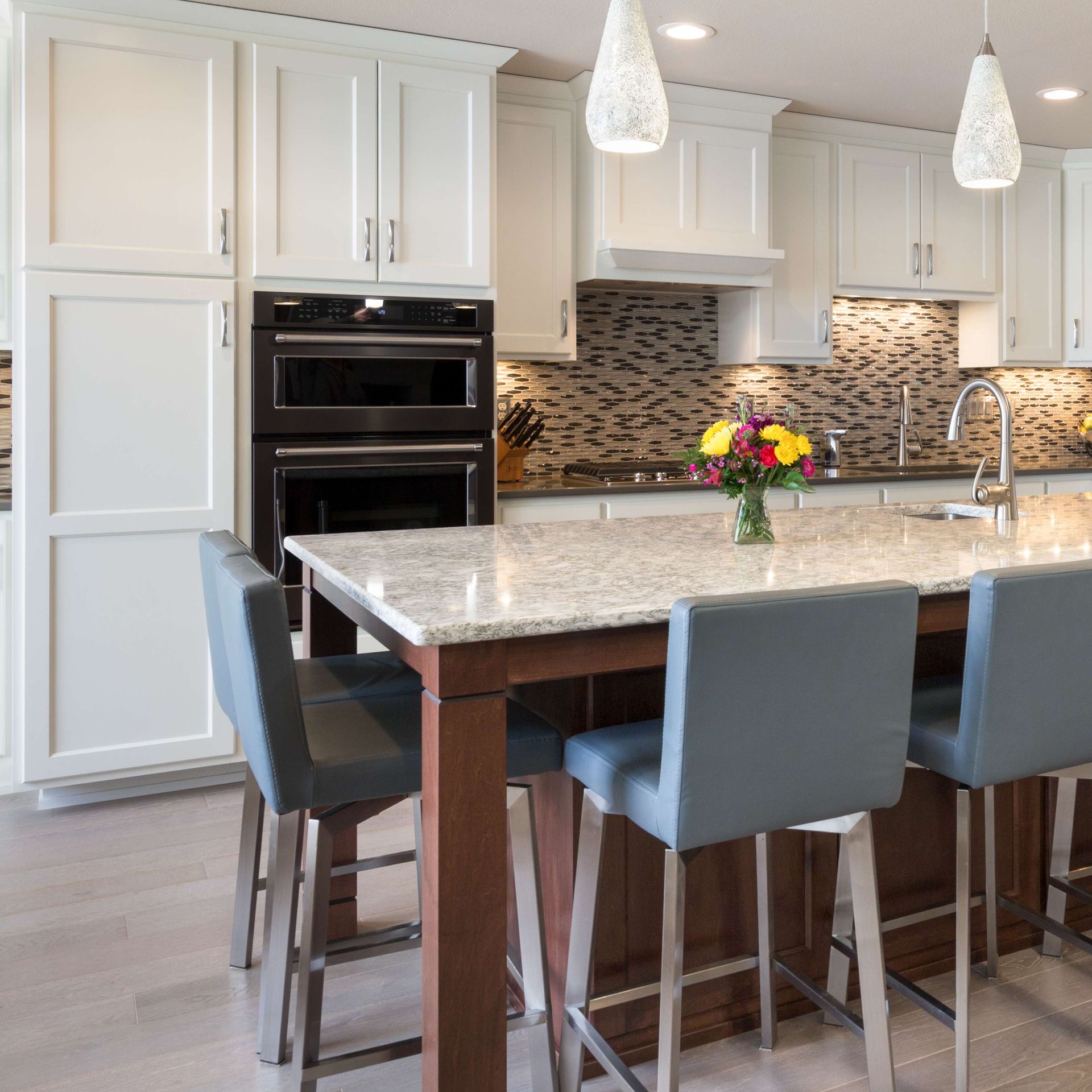 White kitchen with island, gray stools, wood cabinets, black appliances, and pendant lights.