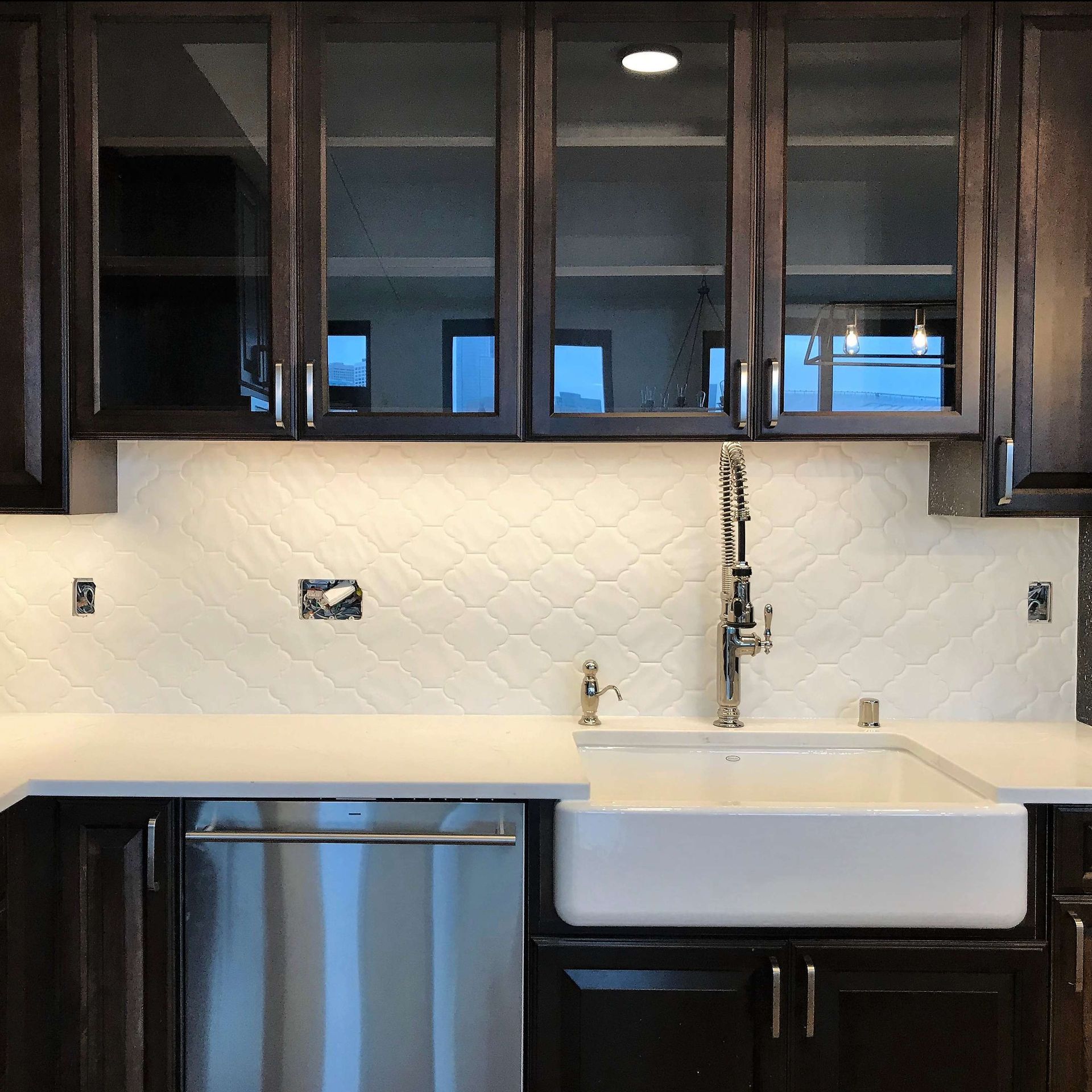 Kitchen with dark cabinets, glass-front uppers, white patterned backsplash, white sink, and stainless steel dishwasher.