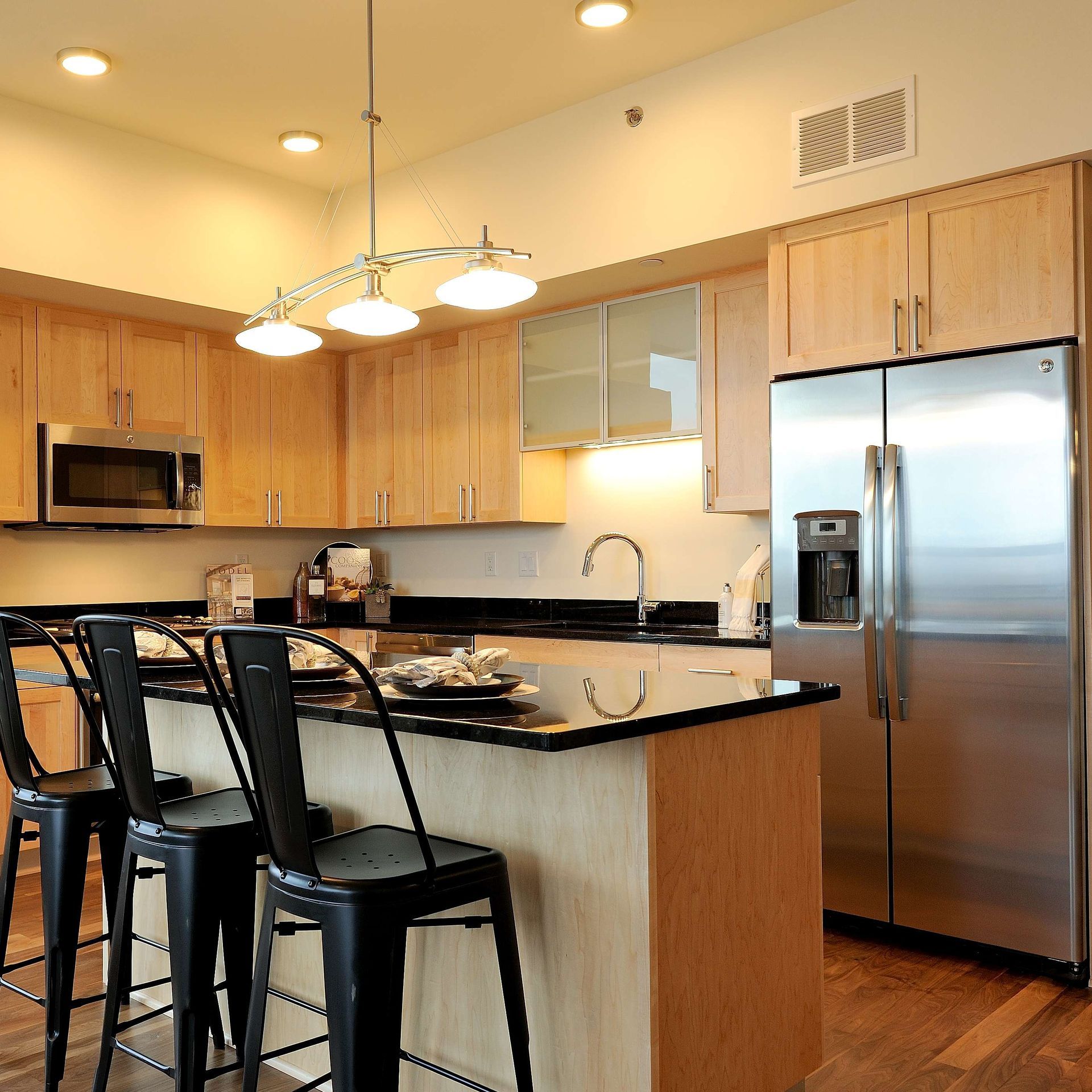 Kitchen with light wood cabinets, stainless steel appliances, and a black countertop island with stools.