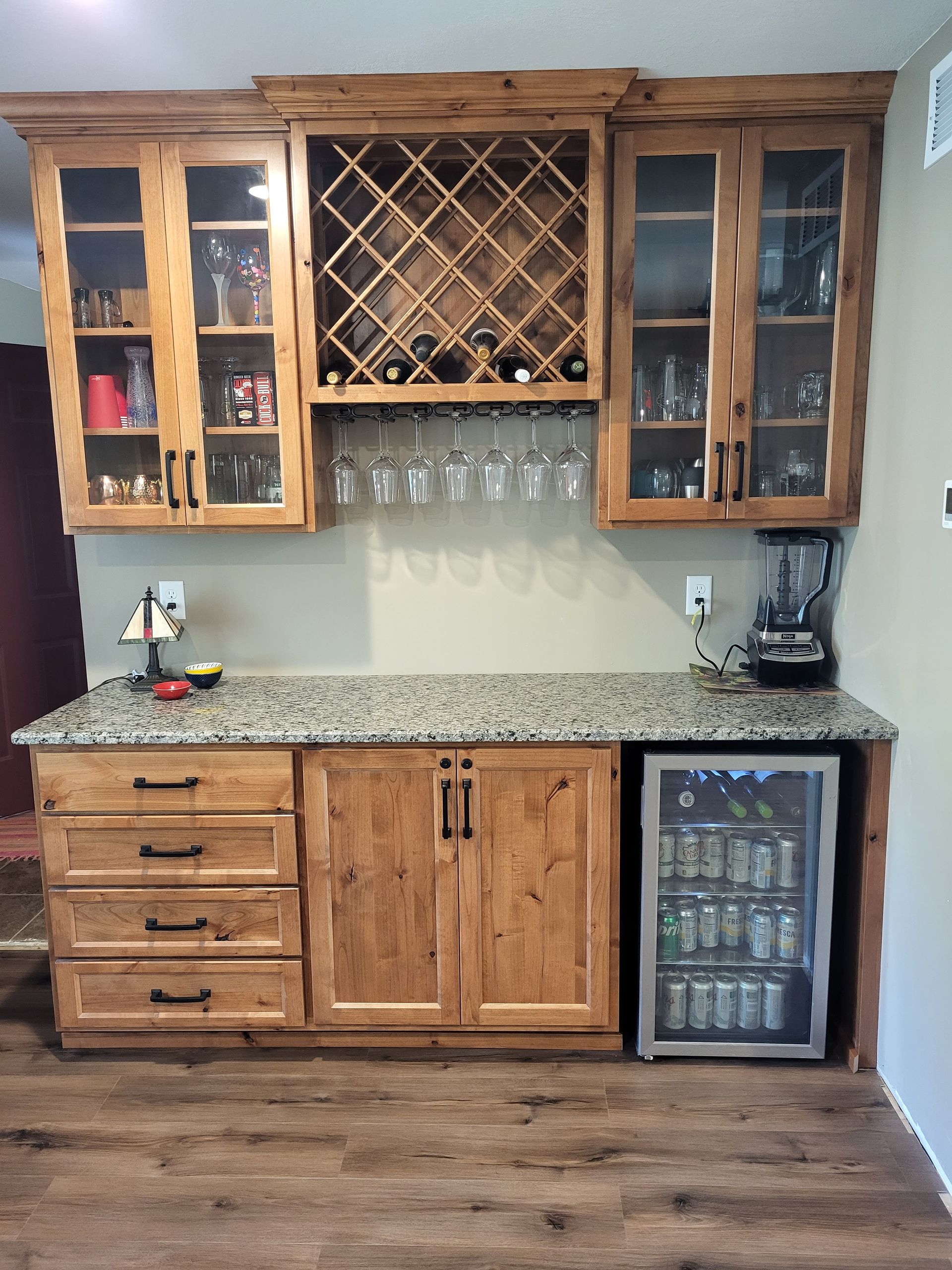 Wooden home bar with upper cabinets, wine rack, glassware, and lower cabinets with granite countertop, mini-fridge.