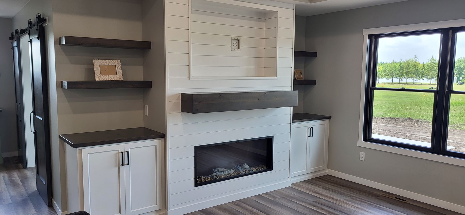 Living room with fireplace, built-in shelves, white cabinets, and a window overlooking a green field.