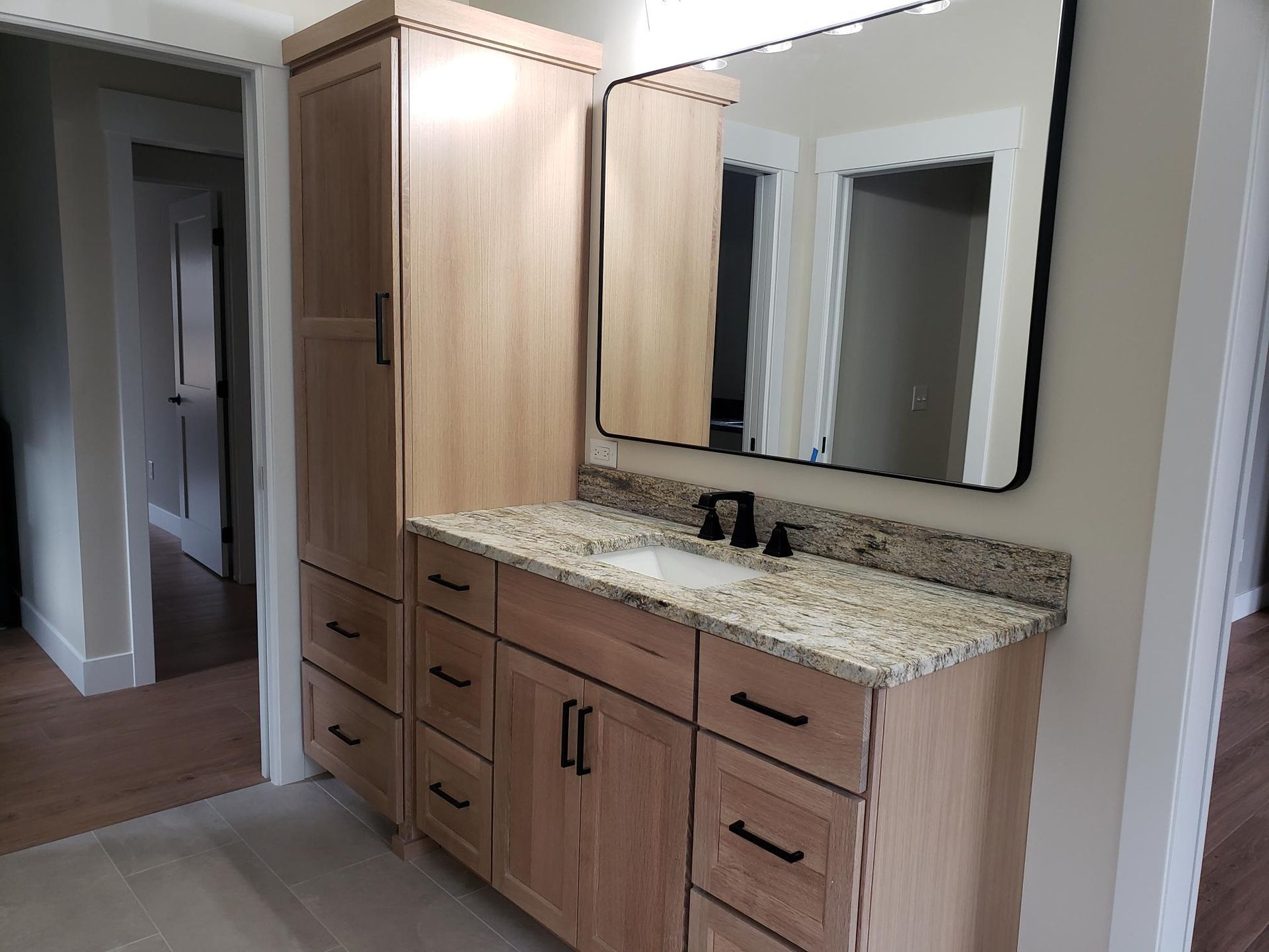 Bathroom with light wood cabinets, granite countertop, black mirror frame, and a tall storage cabinet.