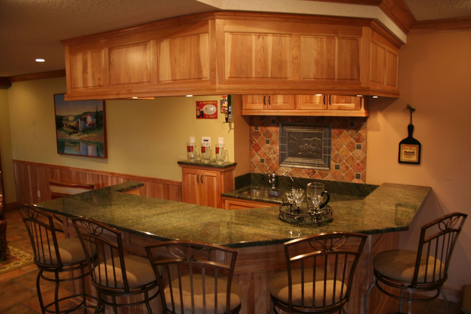 A home bar with a granite countertop, wooden cabinets, and bar stools.  The bar has a tile backsplash and recessed lighting.
