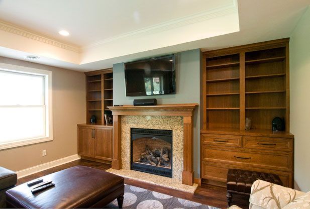 Living room with fireplace, built-in bookshelves, and television mounted above. Brown furniture, neutral walls.