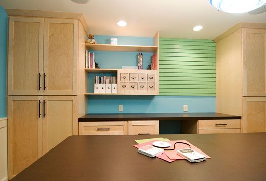 Craft room with light wood cabinets, blue wall, desk, and large table with craft supplies.
