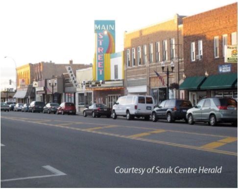 Main Street of a town, buildings with businesses, cars parked along the street.