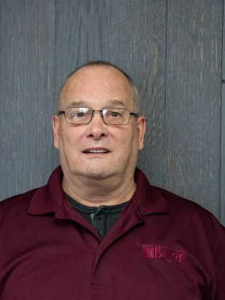 Man wearing glasses and a maroon polo shirt smiles in front of a gray wooden wall.