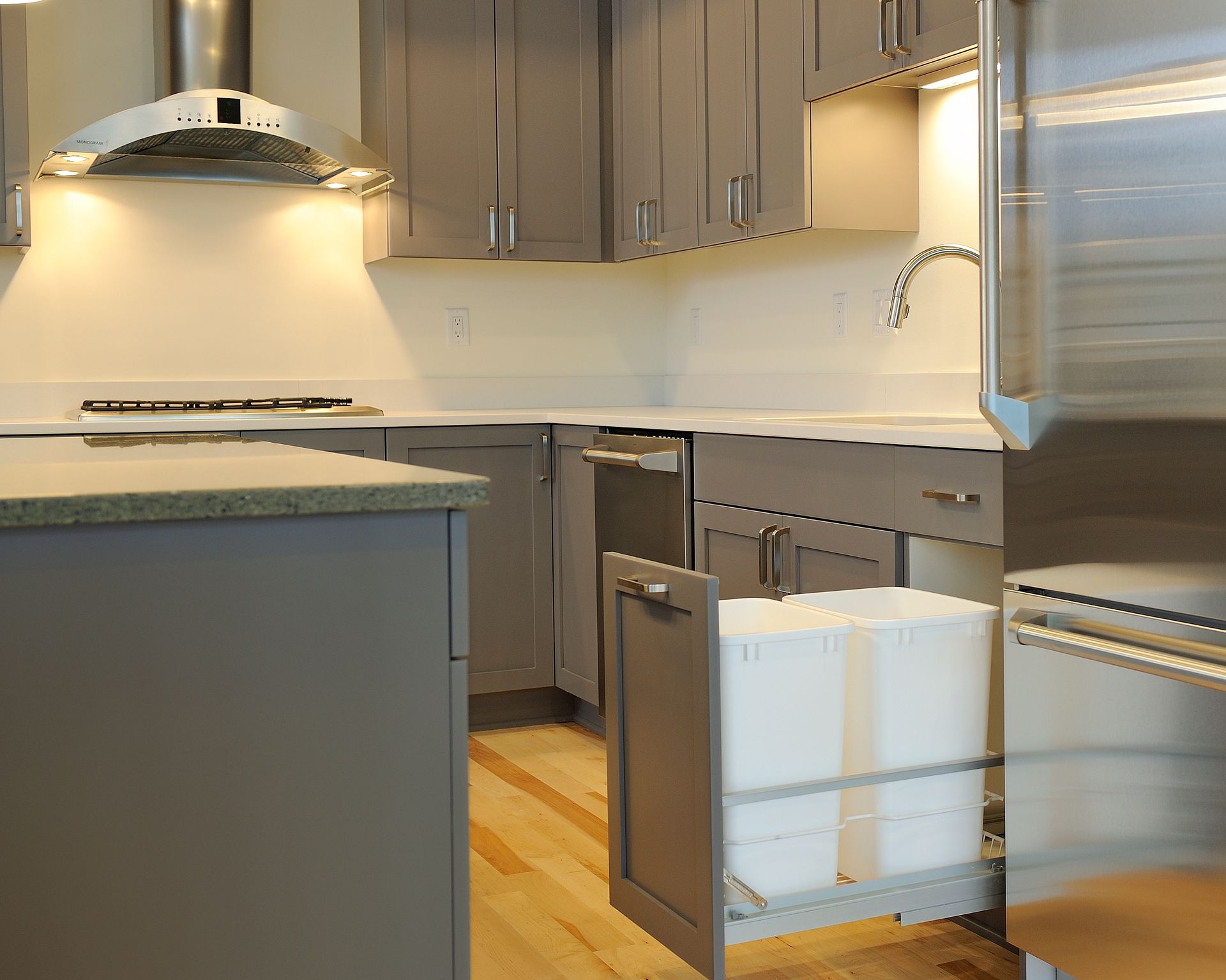 Gray kitchen with pull-out trash bins, stainless steel appliances, and light wood flooring.