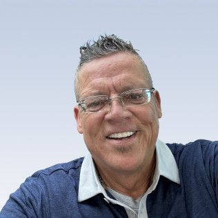 Smiling man with glasses, wearing a blue shirt with a white collar, and a spiky hairstyle.