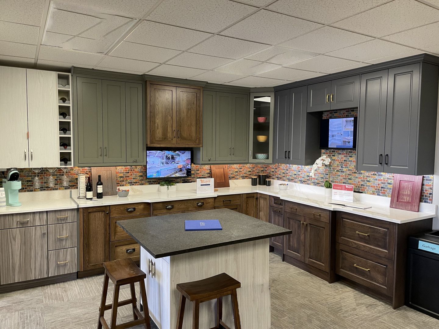 Kitchen cabinet display with various wood tones and countertops, and a small island with stools.
