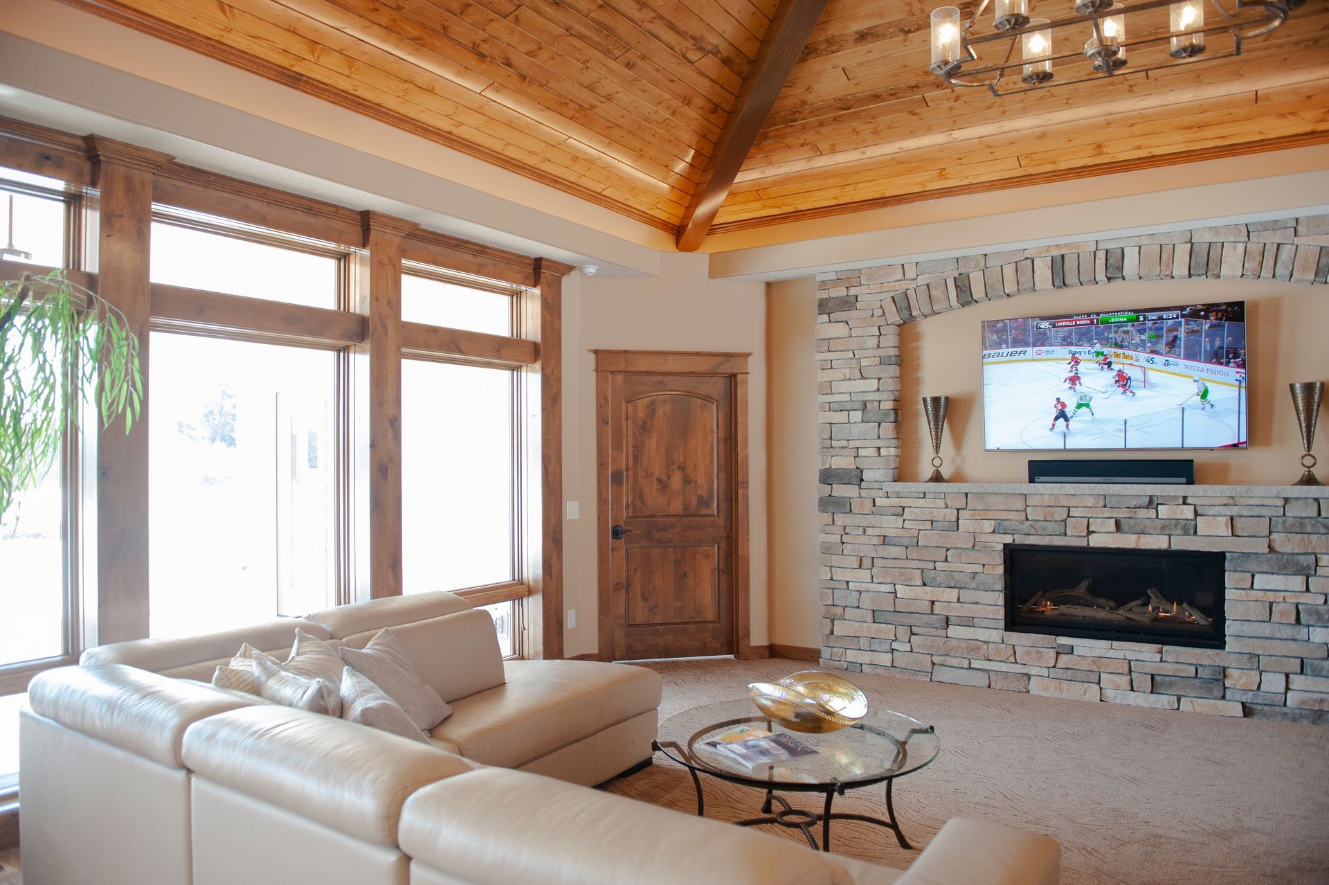 Living room with stone fireplace, TV, sectional sofa, large windows, and wood ceiling.