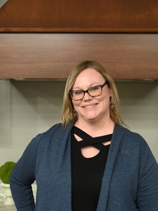 Woman in glasses and blue cardigan smiles, standing in a kitchen with a wooden hood.