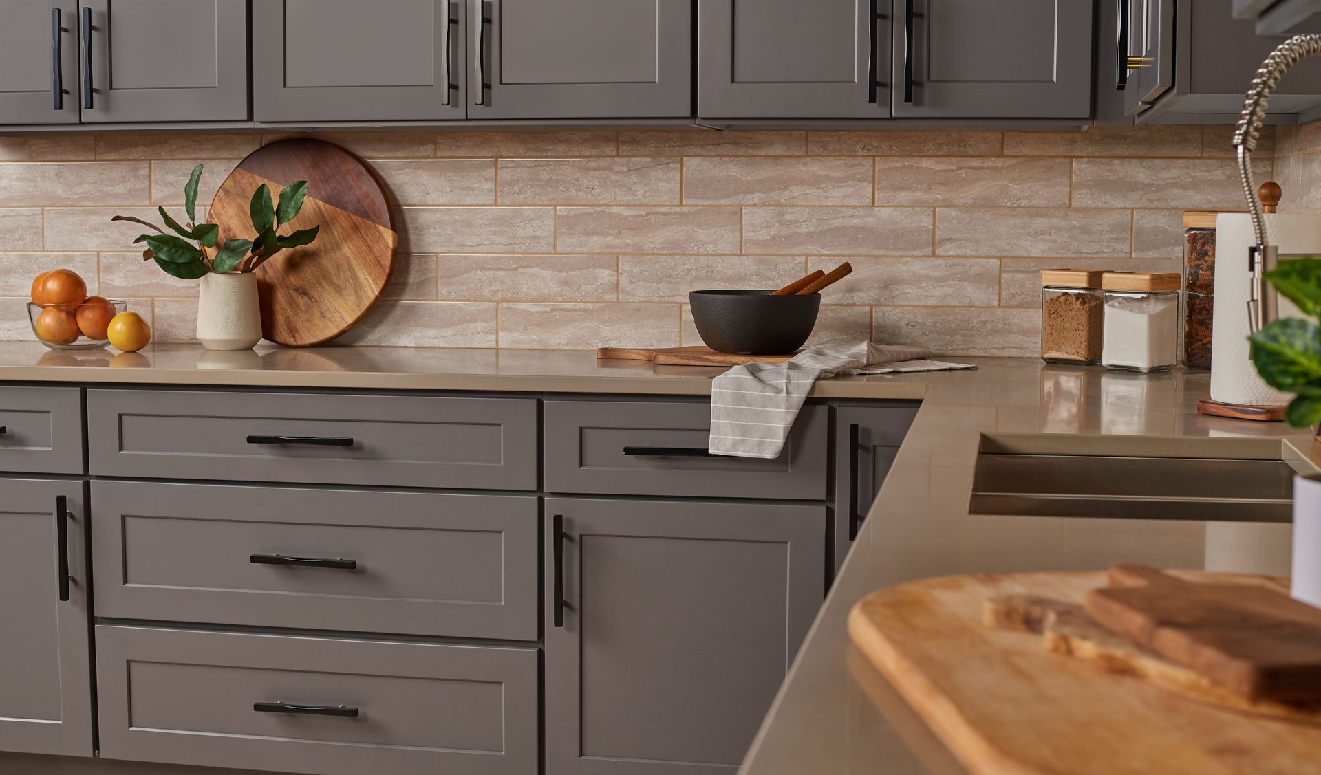 Gray kitchen cabinets with black handles, light-colored backsplash, wooden cutting board, and other kitchen items.