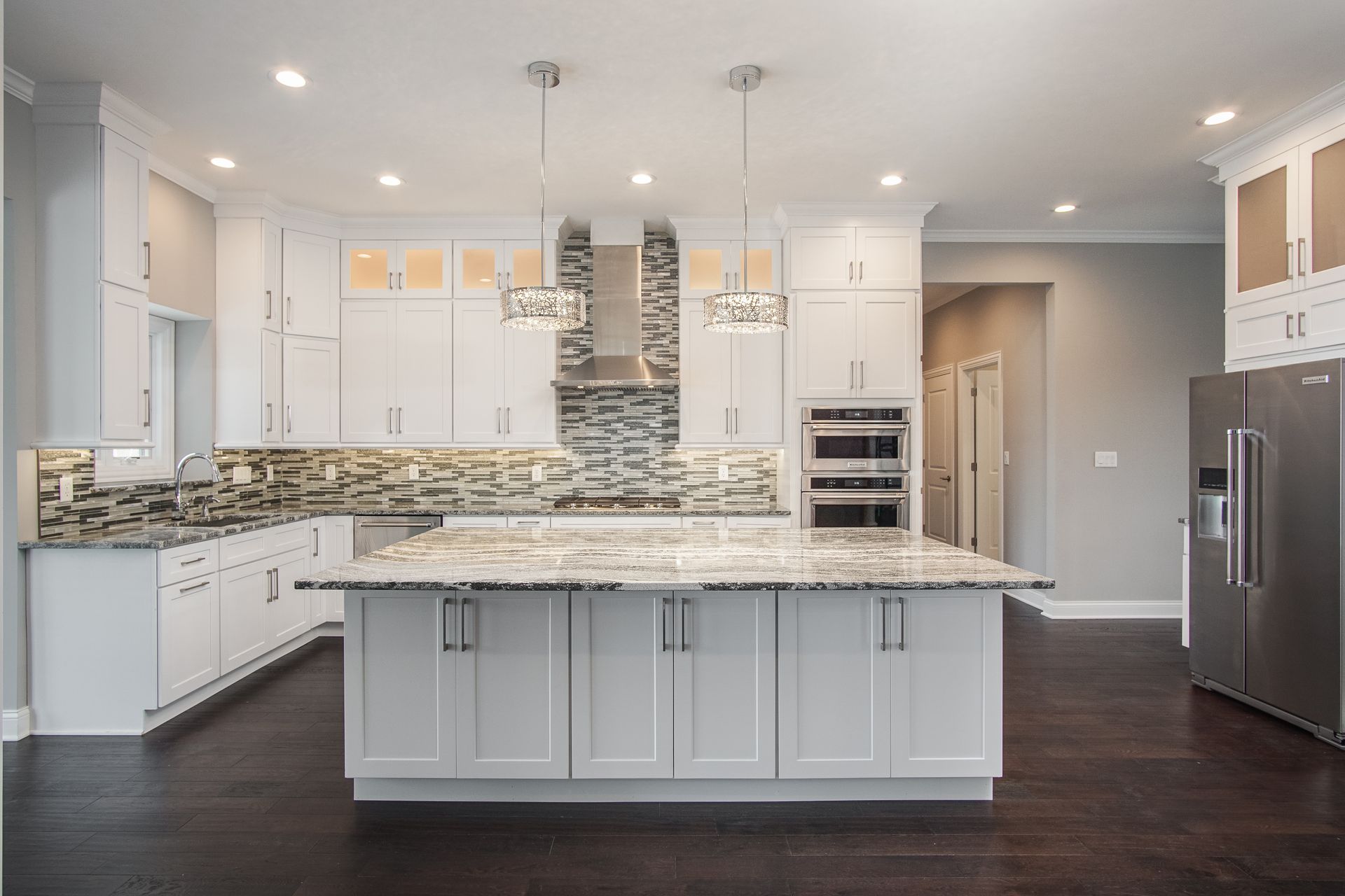 Modern white kitchen with island, dark wood floors, stainless steel appliances, and pendant lights.