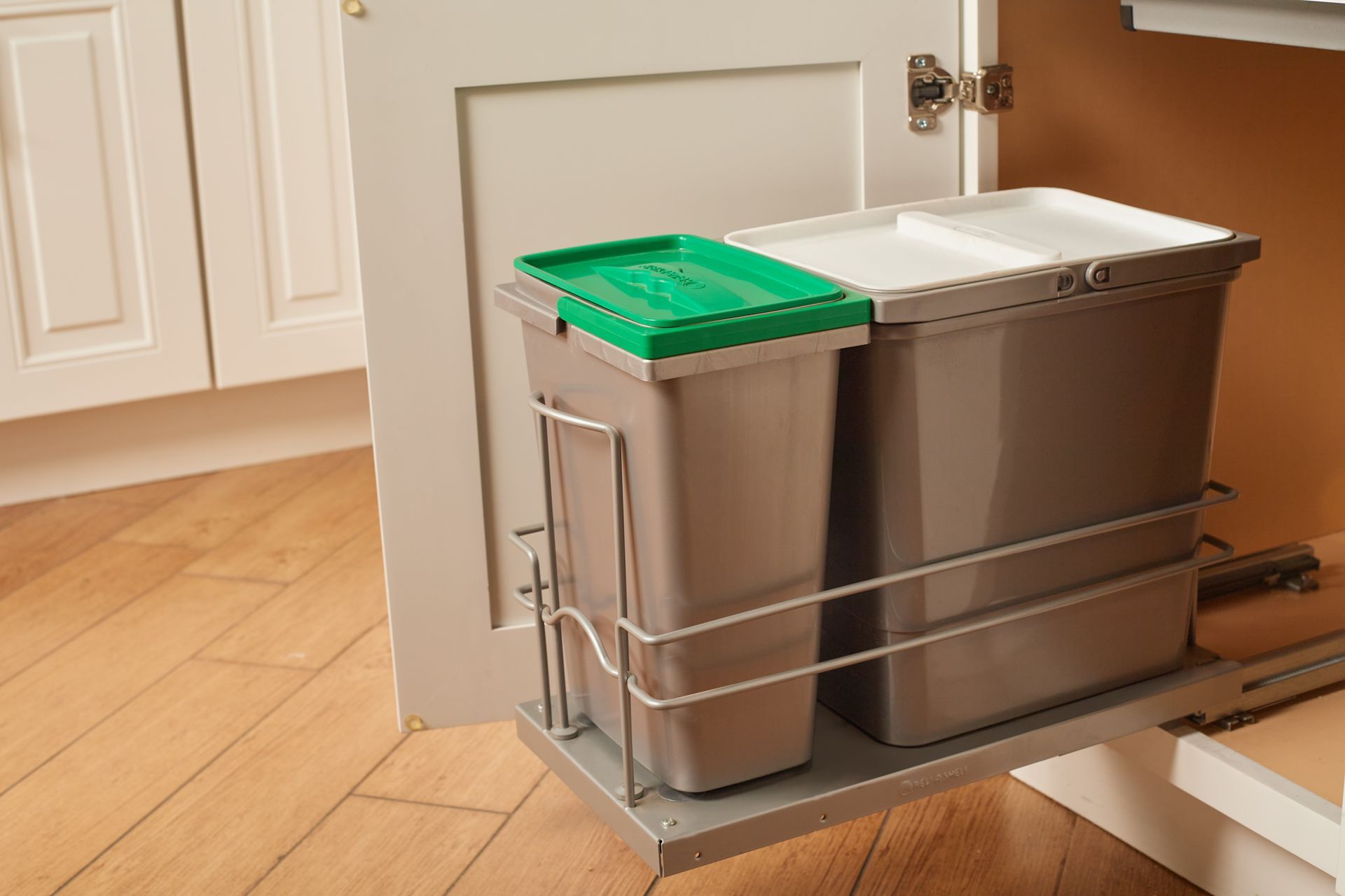 Pull-out kitchen cabinet with two trash bins: one green-lidded, one white-lidded, inside a light gray cabinet.