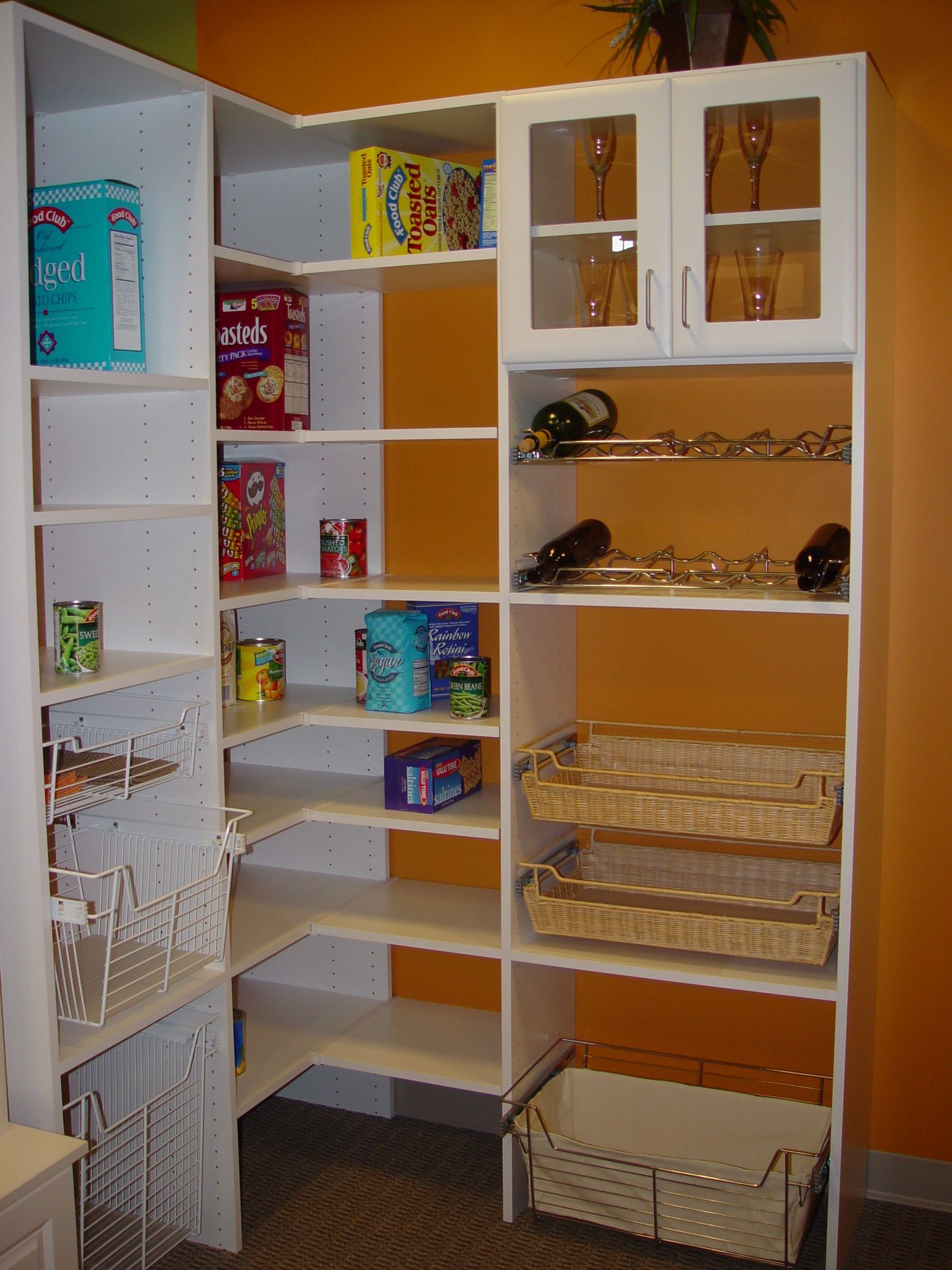 White corner pantry cabinet with shelves and baskets, stocked with food items and glassware.