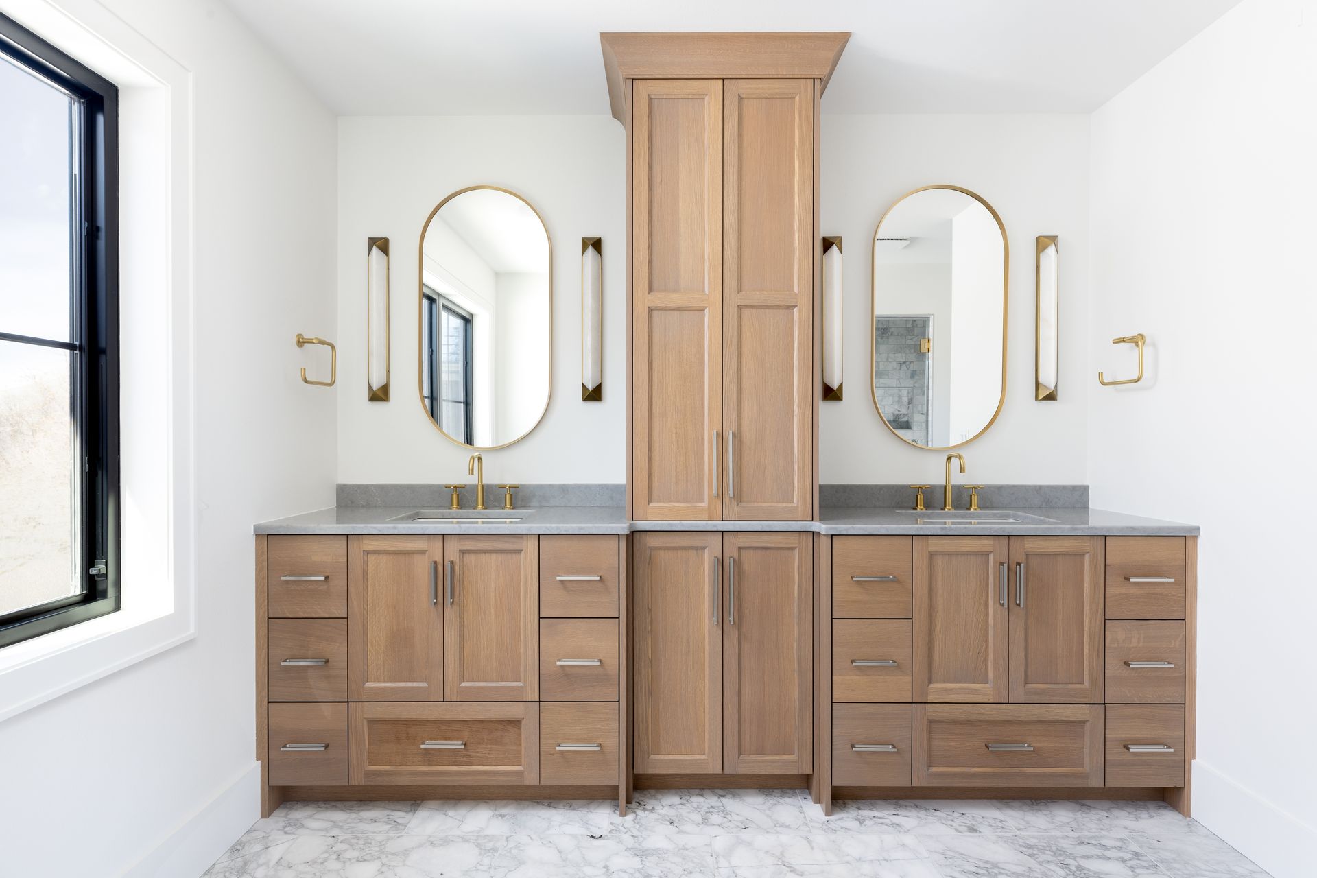 Bathroom with two vanities, oval mirrors, gold fixtures, and light wood cabinetry.