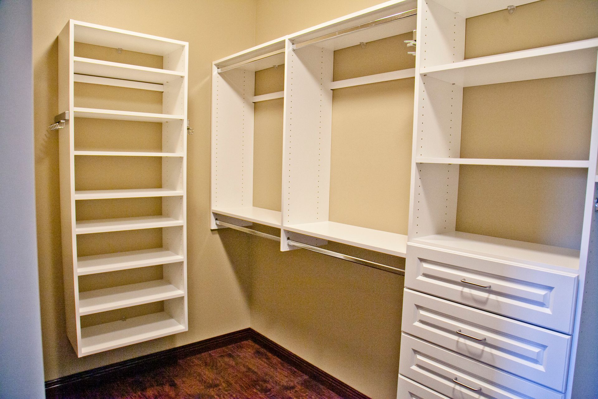 Empty white closet system with shelves, hanging rods, and drawers against tan walls, wooden floor.