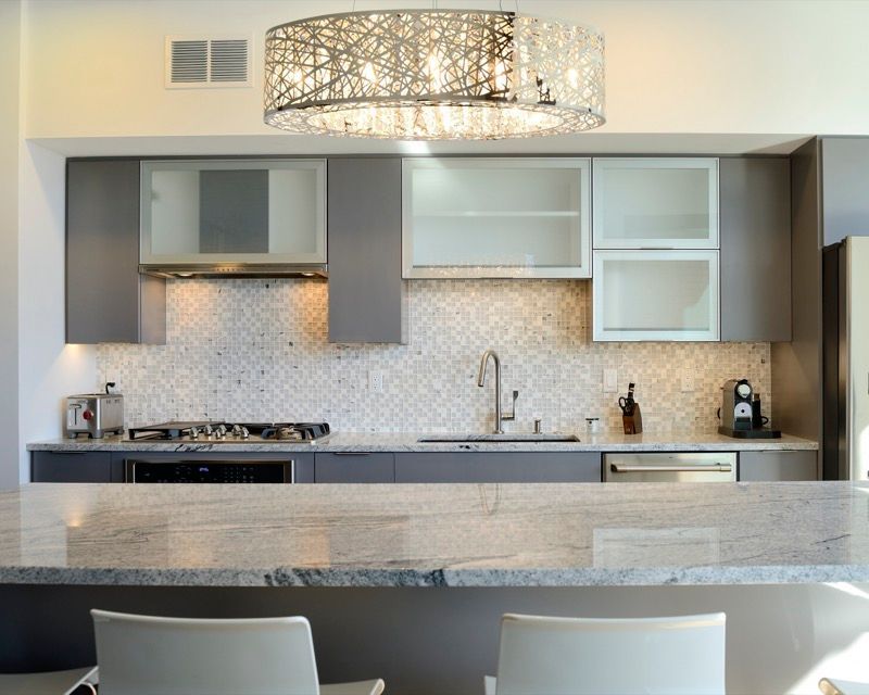Modern kitchen with grey cabinets, mosaic backsplash, and granite countertop, with a chandelier overhead.