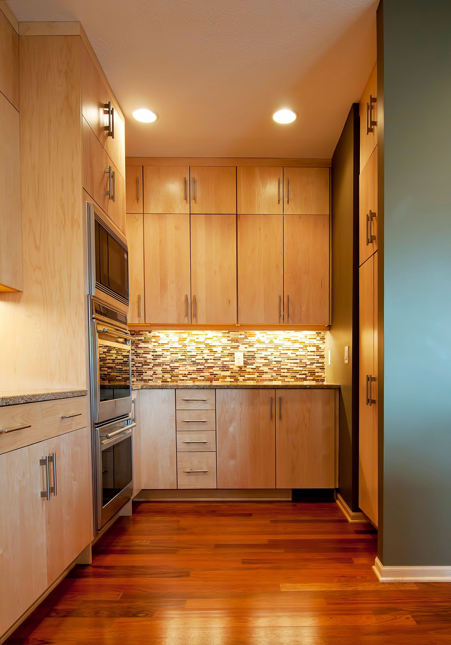 Narrow kitchen with light wood cabinets, stainless steel appliances, and wood flooring.