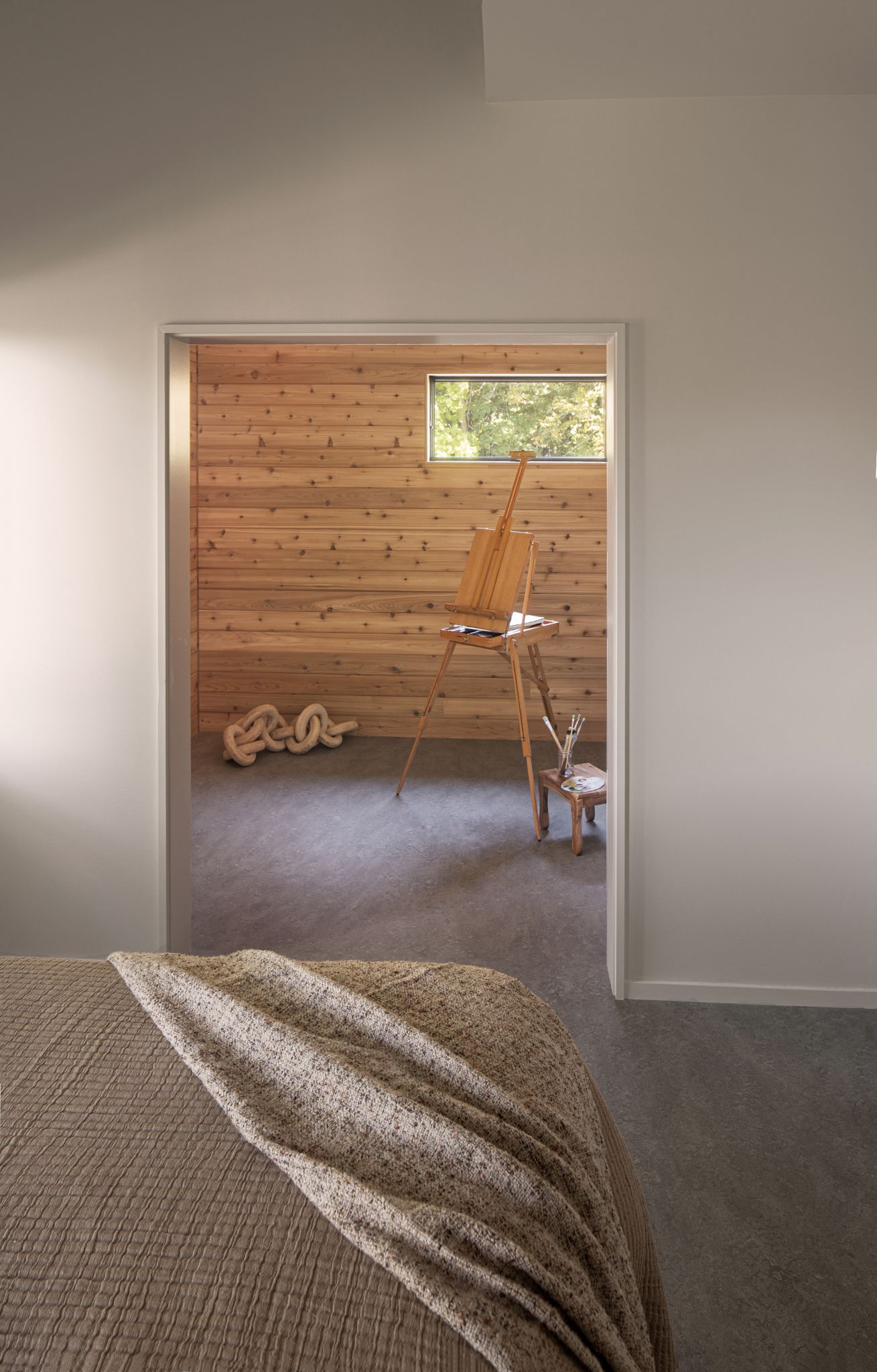 View from a room with a bed toward a wood-paneled room with an easel and window.