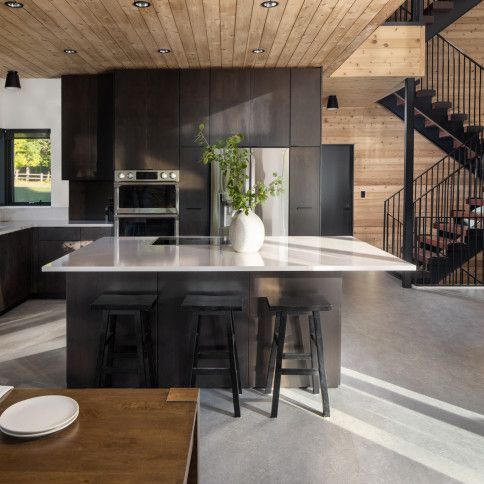 Modern kitchen with dark cabinetry, white countertop island, wooden ceiling, and stairs.