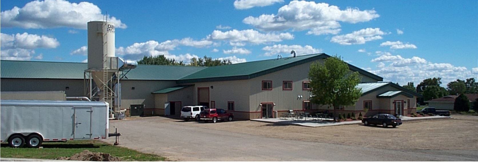 Large industrial building with silo, vehicles, and a trailer on a bright sunny day.