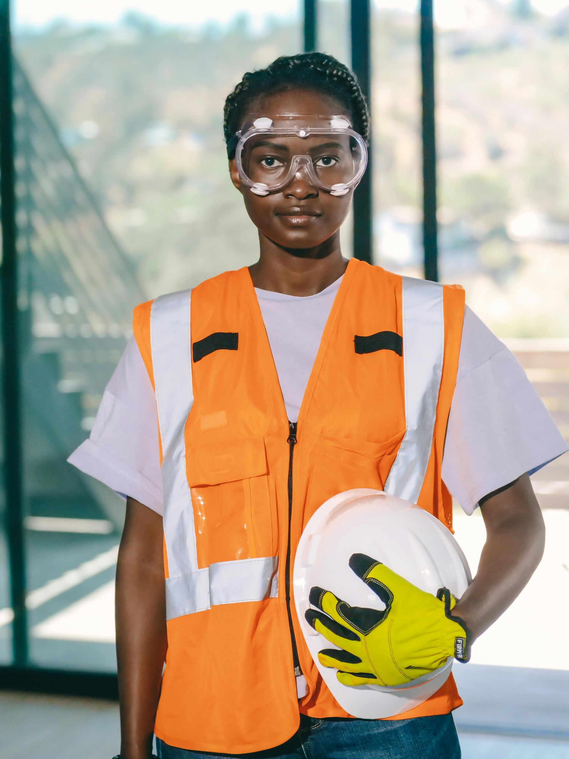 Woman in safety gear, holding a hard hat. Wearing orange vest, safety glasses, and gloves. Indoors, construction setting.