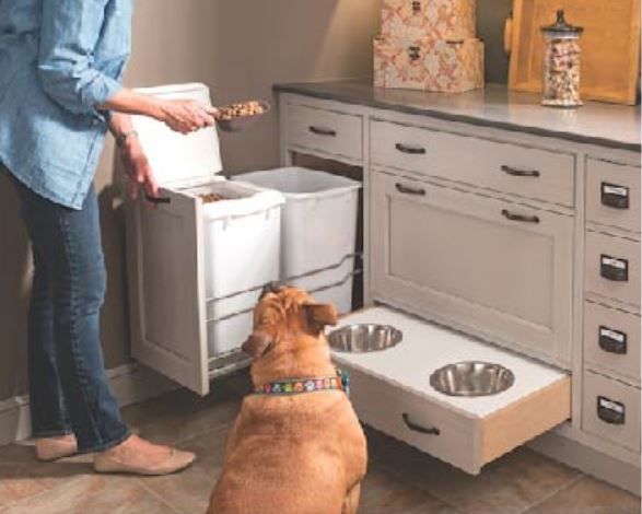 Woman filling dog food dispenser. Dog looks up expectantly. Built-in dog bowl and storage in a kitchen.