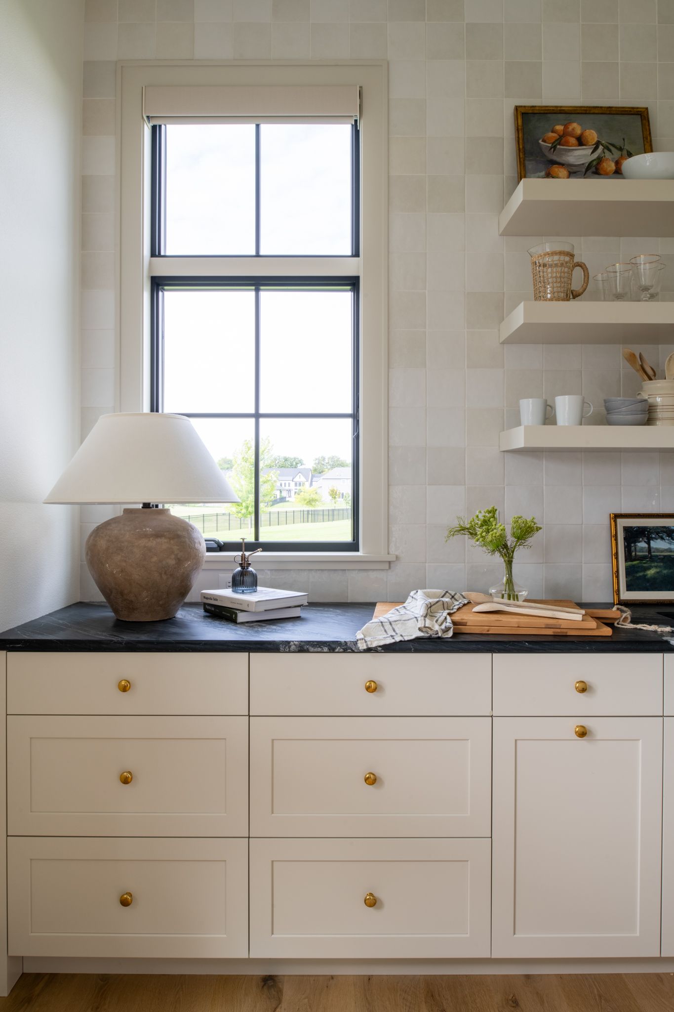 Cream-colored kitchen cabinets with brass knobs, black countertop, a window with black trim, and floating shelves.