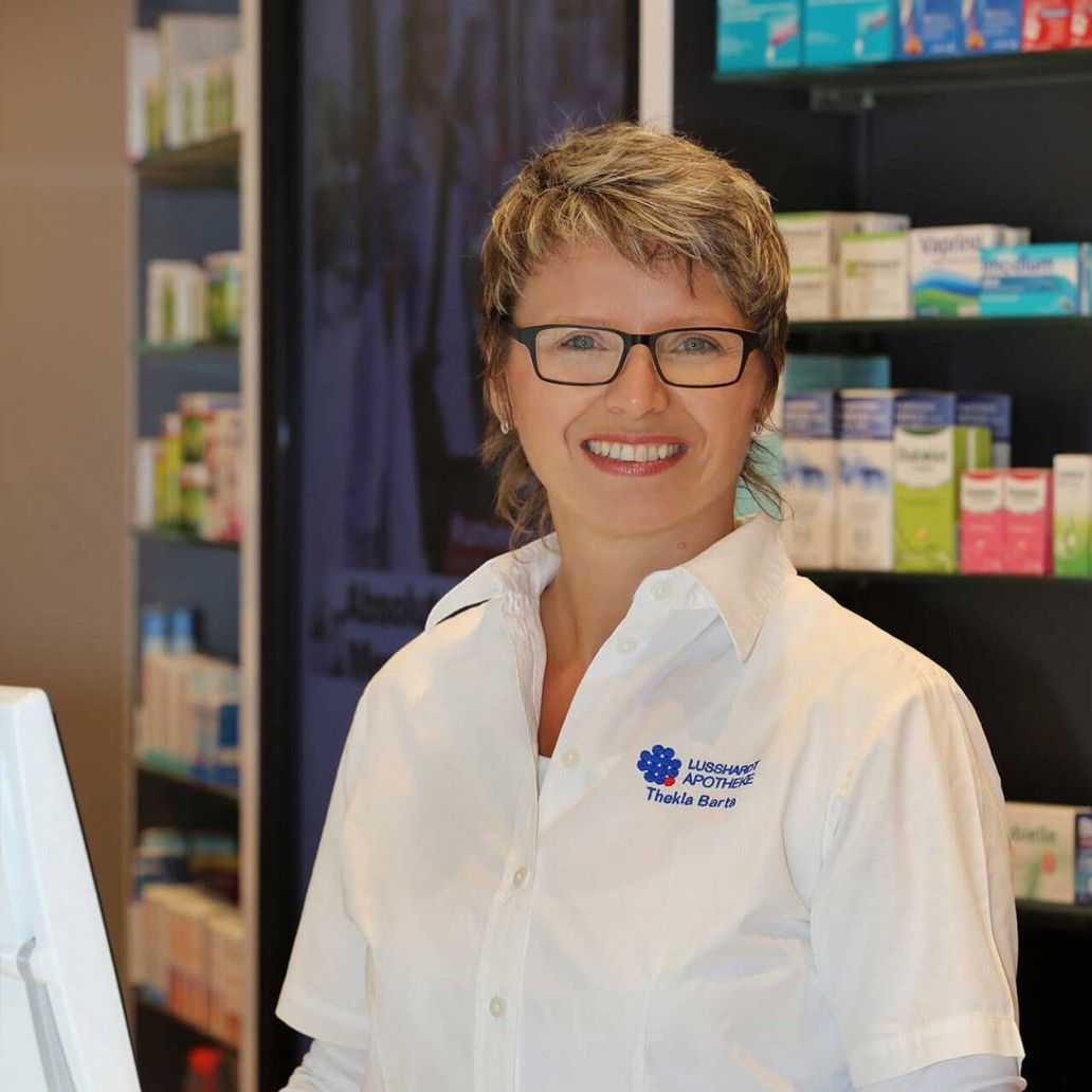 Eine Frau mit Brille und weißem Hemd lächelt in einer Apotheke