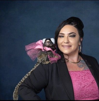 Woman in pink top and black blazer, with monkey wearing a pink tutu on her shoulder, dark blue background Woman in pink top and black blazer, with monkey wearing a pink tutu on her shoulder, dark blue background