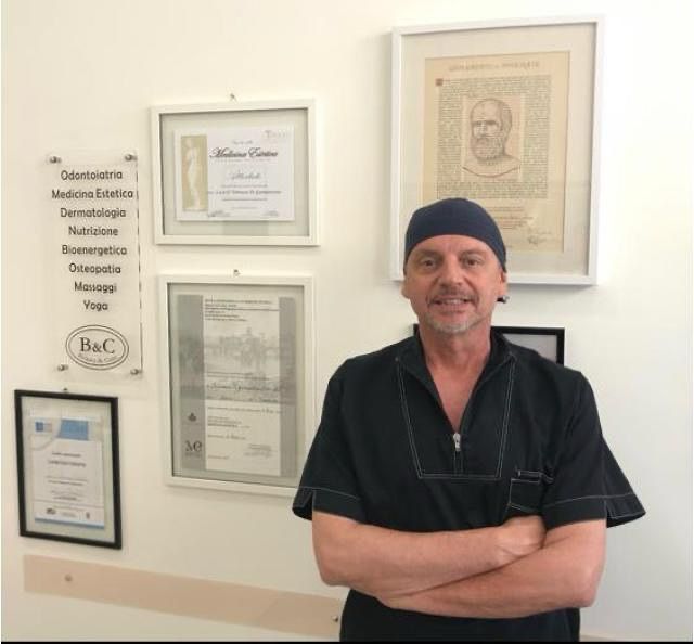 Un uomo con le braccia incrociate è in piedi davanti a documenti incorniciati, tra cui certificazioni, in un ambiente clinico.