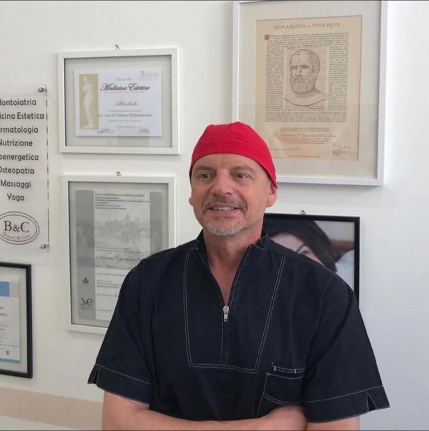 Un medico con le braccia incrociate, che indossa un berretto blu e un camice bianco, è in piedi davanti a dei certificati incorniciati.