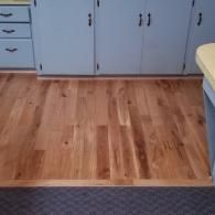 Light blue kitchen cabinets above a natural wood-plank floor, bordering a dark gray carpeted area.