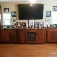 A large flat-screen TV mounted above a long wooden cabinet cluttered with numerous framed photographs and a window.