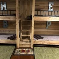 Wooden bunk beds for four with a central ladder, featuring metal letters H, B, E, and P mounted on the rustic wood walls.