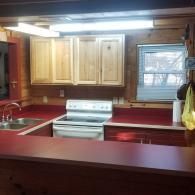 A rustic-style kitchen with natural wood cabinets, red countertops, a white electric stove, and a window above the sink.