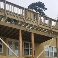 A tan two-story house with a wooden deck featuring a railing, supported by upright wooden posts and diagonal braces.