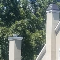 Two stucco chimneys against a backdrop of lush green trees, one featuring a small metal vent and the other a chimney cap.