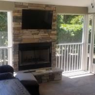 A stone fireplace with a mounted television in a screened-in porch, featuring white railings and views of outdoor greenery.
