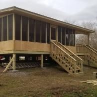A newly constructed wooden cabin on stilts with a screened-in porch and wooden stairs leading to the ground.