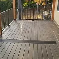 A deck with grey-brown wooden planks and a dark-toned accent board, featuring a black metal railing overlooking trees.