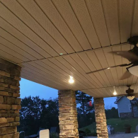 A covered outdoor patio featuring stone columns, a beige ceiling with recessed lights, and two ceiling fans.