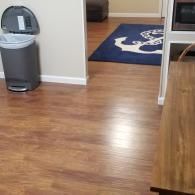 A gray pedal trash can sits on a wood-look floor next to a doorway leading to a room with a blue anchor-patterned rug.