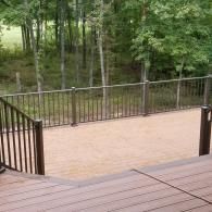 A wood-tone composite deck featuring brown metal railings and a staircase, overlooking a lush green forest.
