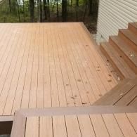 A light brown wooden deck with a staircase leading up to a beige-sided house, surrounded by trees.