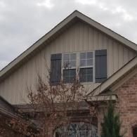 A triangular gable with tan vertical siding, a central window with dark shutters, and a partial brick wall below.