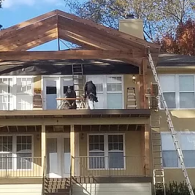 Two workers install a wooden roof frame on the upper deck of a multi-story house. A tall ladder leans against the side.