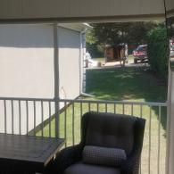 A shaded porch with a comfortable armchair and side table overlooking a sunny backyard with a lawn and a parked truck.